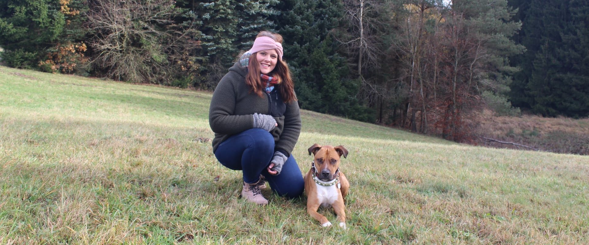 Frau sitzt in der Hocke, brauner Hund liegt neben ihr auf der Wiese